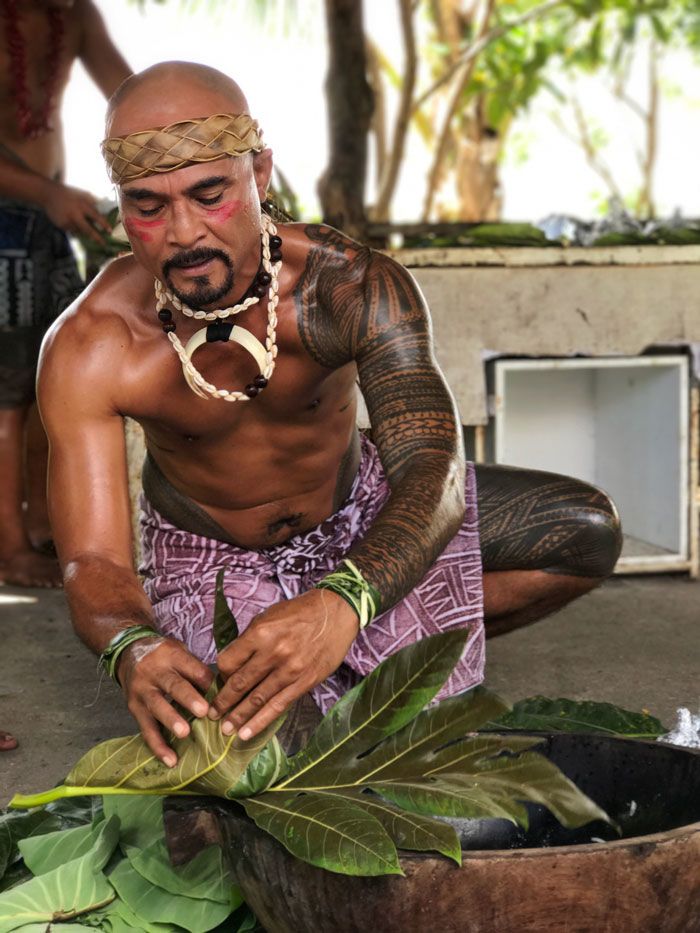 A heavily tattooed Samoan man wearing traditional jewelry and purple fabric draped around his waist wraps leaves around food.