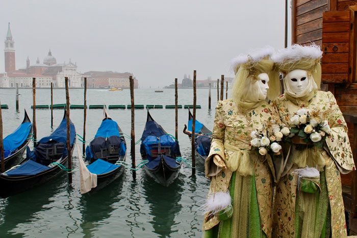 Two people wearing blank white masks and ornate yellow and green dresses hold white roses while standing by water where small boats are tied up in Italy.