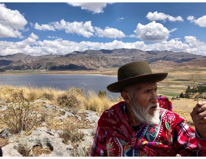 A Peruvian man wearing a hat and brightly colored clothing stands in the foreground with a lake and mountains behind him.
