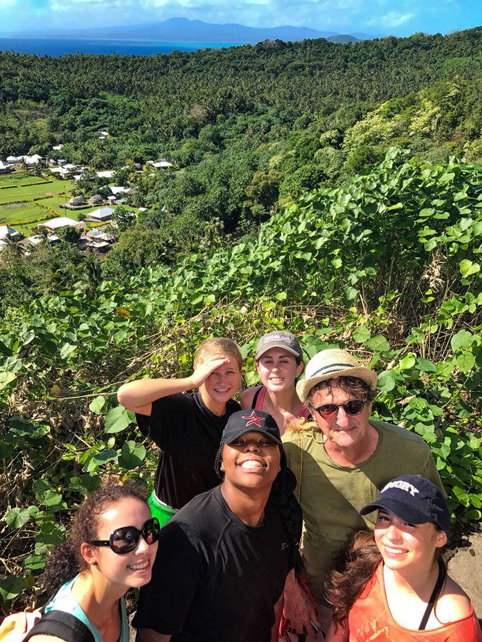 Students pose for a selfie in Samoa with lush trees and mountains behind them.