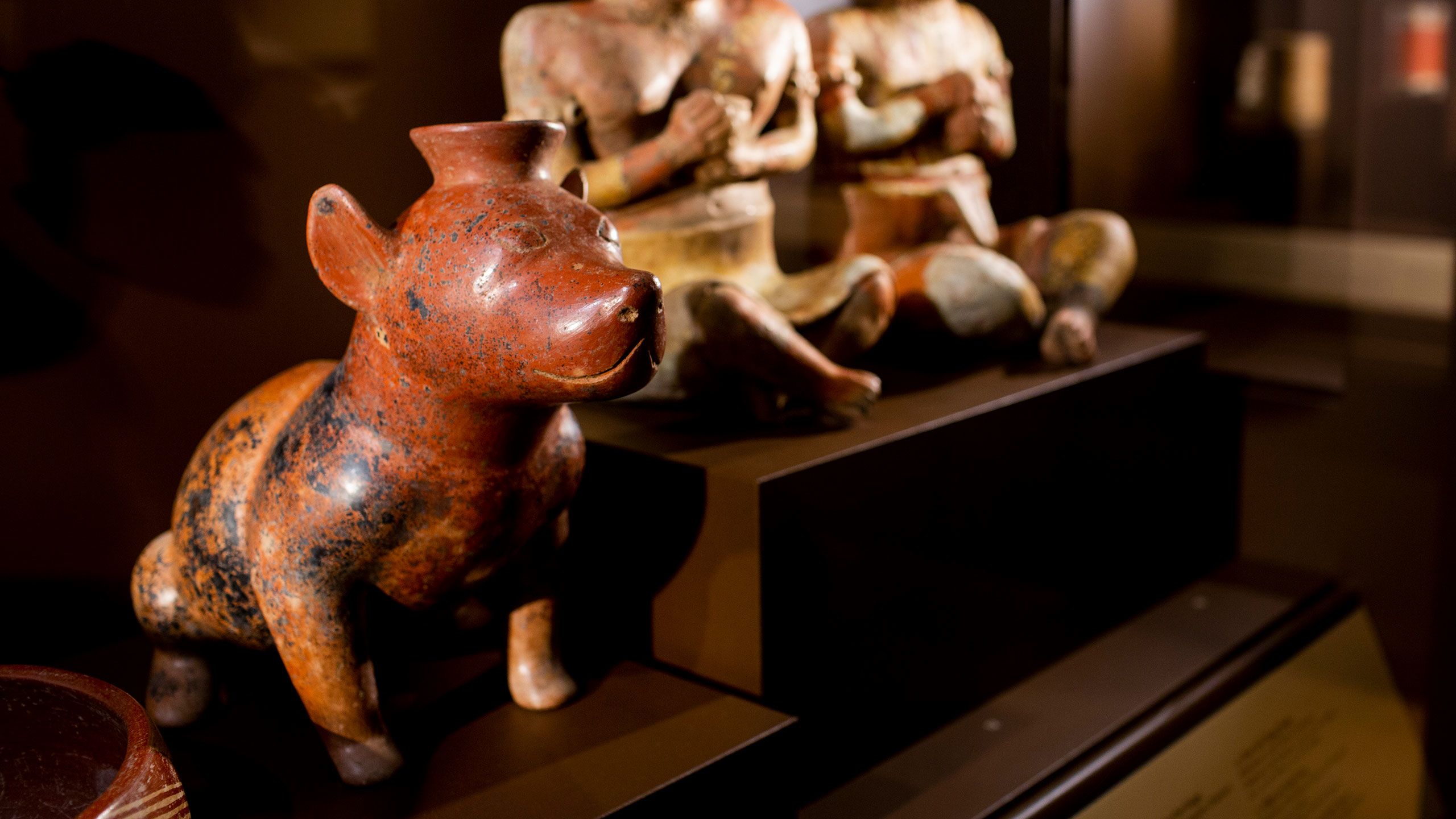 A small ceramic orange-brown dog figurine with a mottled surface sits in a display case with two ceramic figurines showing people sitting with legs crossed and arms folded. 