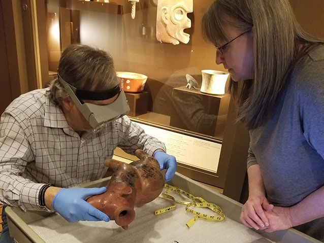 Forensic anthropologist Robert Pickering wears blue gloves and an eyepiece as he examines a ceramic dog in the Carlos Museum. A tape measure lies on the cart next to the object and a woman wearing glasses looks on.