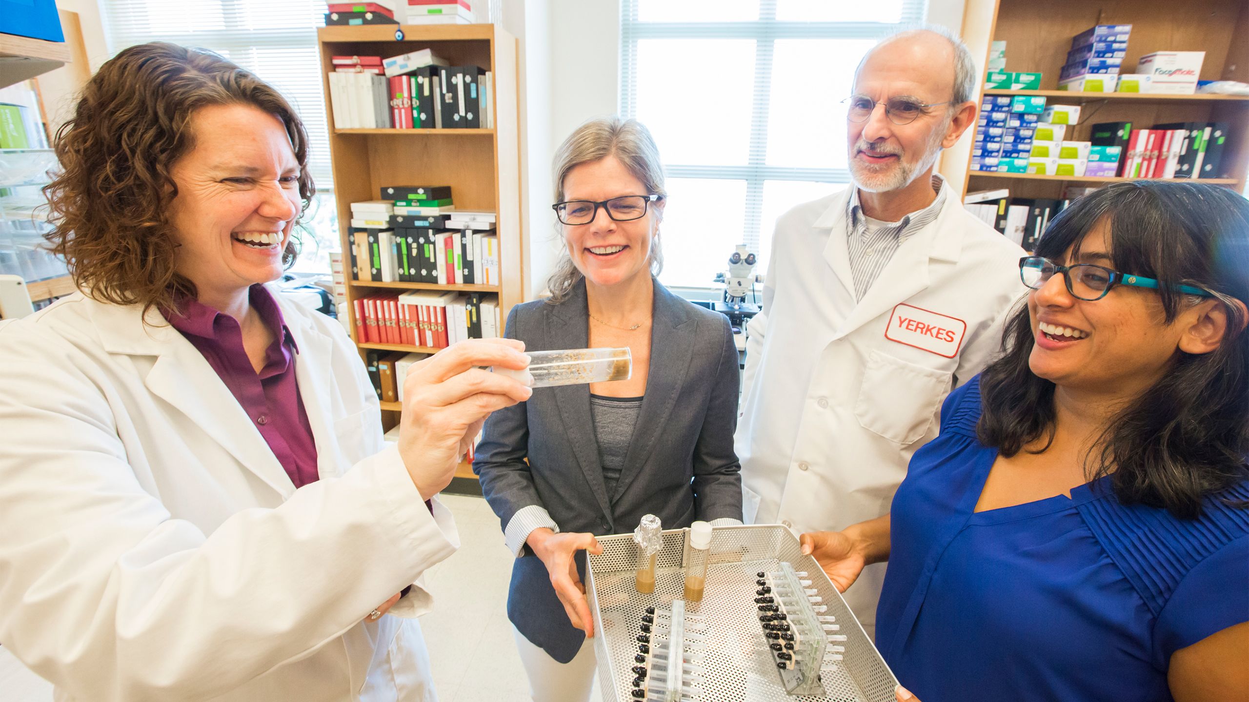 Emory scientists Amanda Freeman, wearing a white lab coat, shows a glass vial to playwright Margaret Baldwin, professor Lary Walker and playwright Natasha Patel.