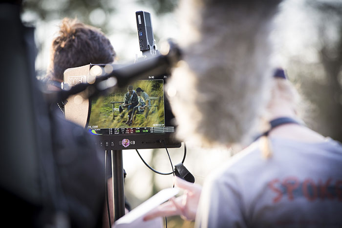 Two Emory students man a camera and watch through a small screen a scene being filmed of two actors talking to each other on a bench. A boom microphone is artfully out of focus in the photo.