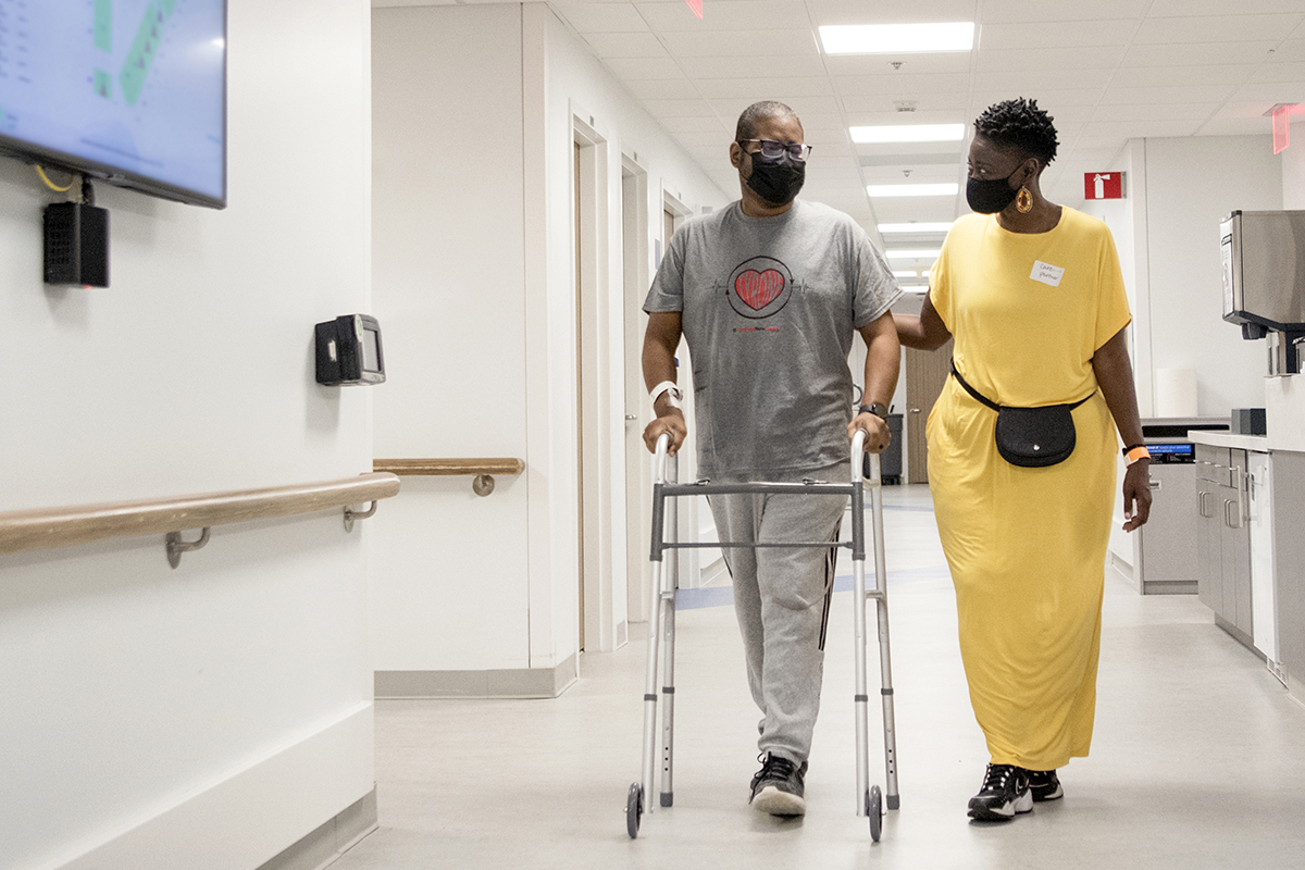 A man wearing a t-shirt with a heart emblem walks in a hospital corridor using a walker. A woman walks beside him holding his arm.