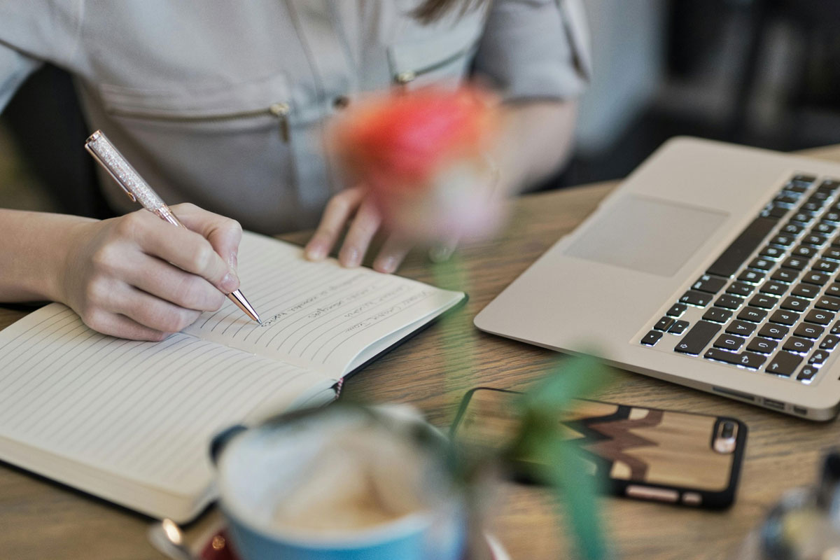 close up view of a person writing in a notebook with a laptop on the table nearby