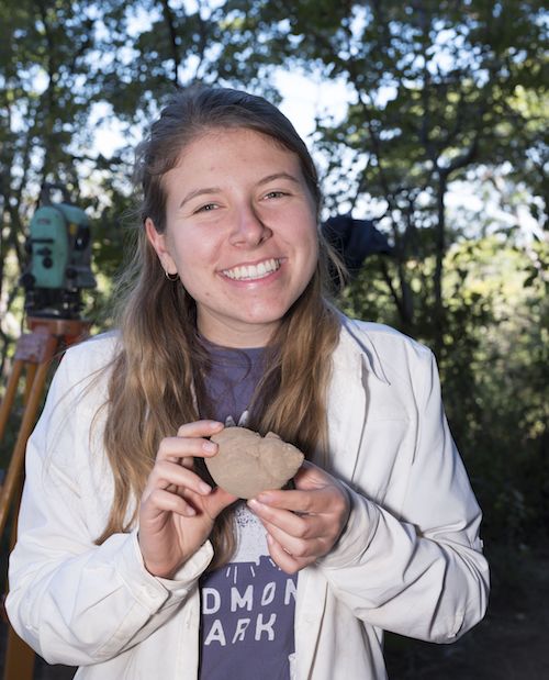 Student Suzanne Kunitz hold an African land snail shell at a dig site in Malawi with trees and equipment in the background.