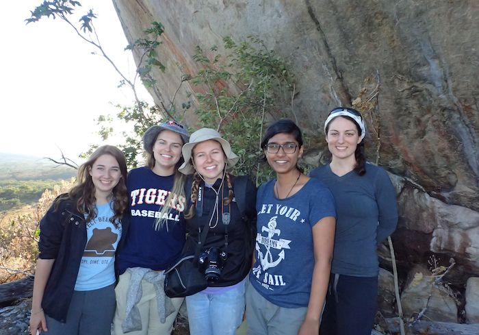Students pose with arms around each other, standing in front of a tall rock surface.