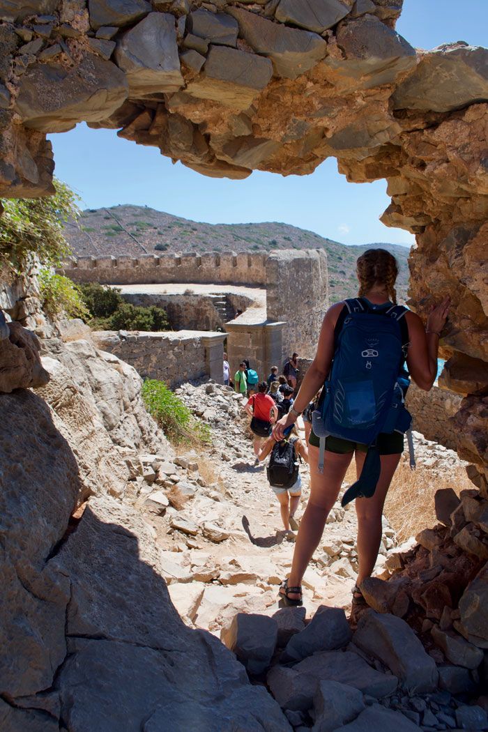 Students wearing shorts and carrying backpacks hike down a rocky trail amid ruins in Greece.