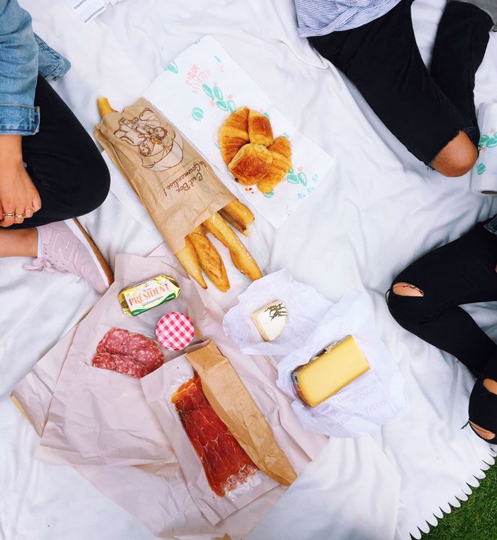 A photo taken from above shows the knees and feet of students sitting on a picnic blanket in France, with cheese, meat, croissants and loaves of bread spread out on the blanket.