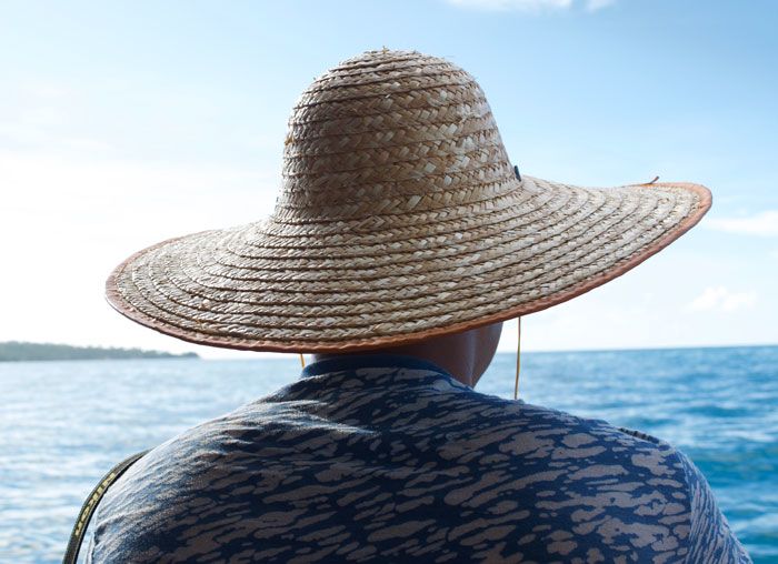 A Samoan man in a large hat is photographed from behind as he looks out at the ocean.