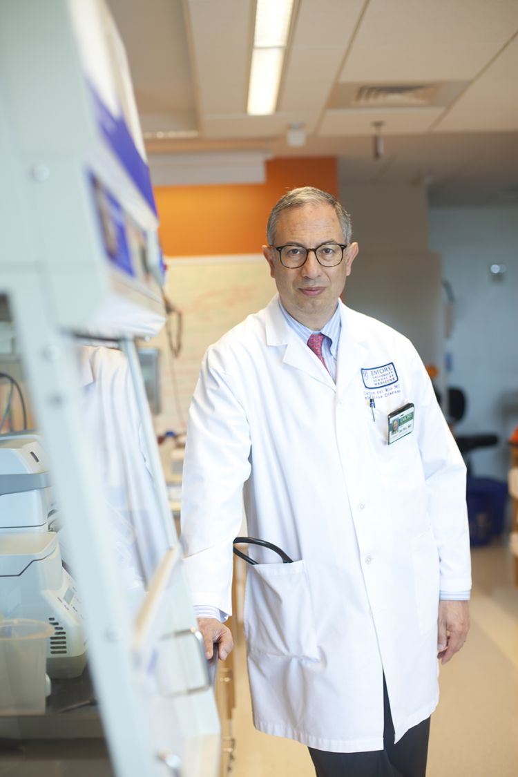 Wearing a white lab coat, Carlos del Rio leans against a panel.