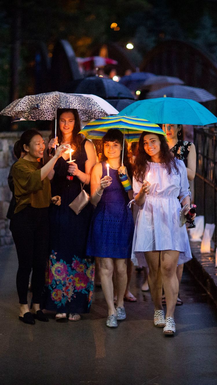 Students carrying umbrellas and lit candles cross the Emory Bridge over Houston Mill Road, which is lined with votive candles in white bags.