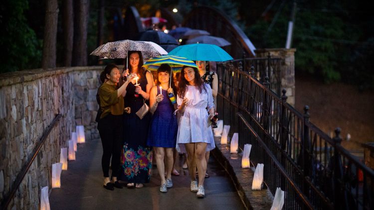 Students carrying umbrellas and lit candles cross the Emory Bridge over Houston Mill Road, which is lined with votive candles in white bags.