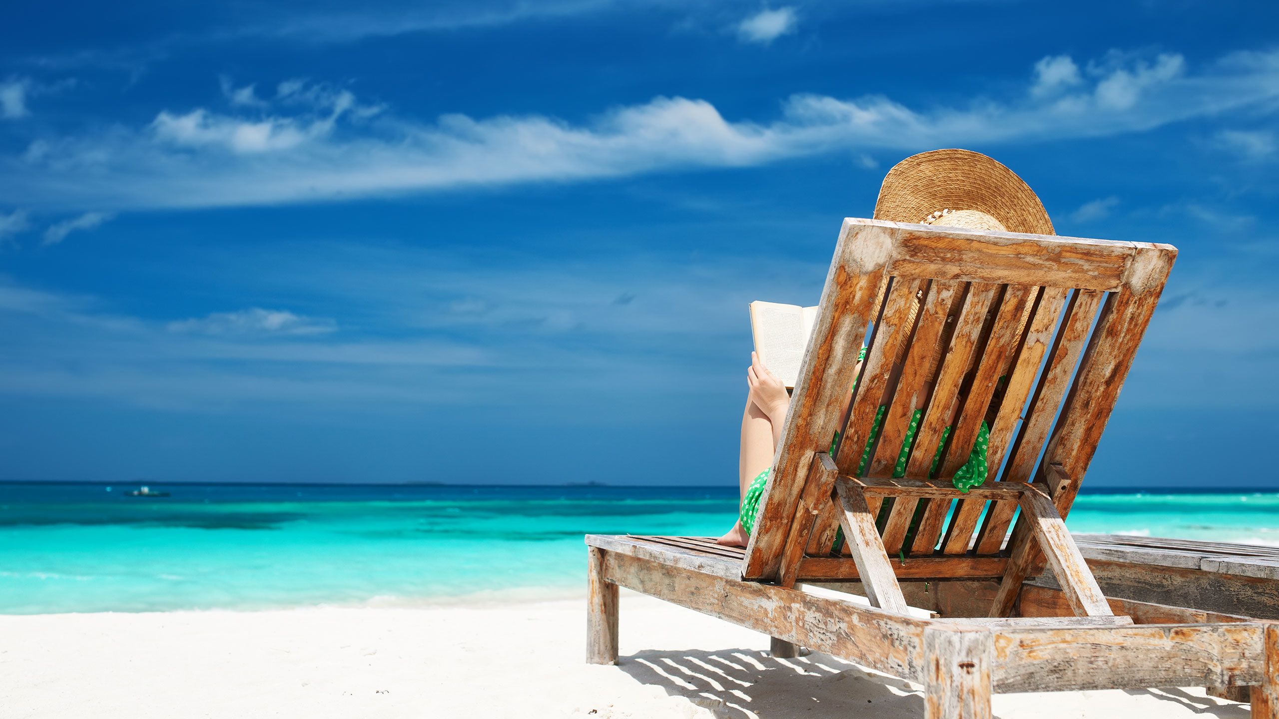 A woman reads a book in a wooden lounge chair on the beach, looking out at the blue ocean and blue sky