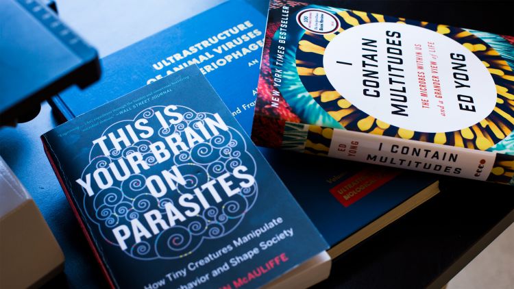 Three books related to the human microbiome sit on a table. The titles are "This is Your Brain on Parasites," "Ultrastructure of Animal Viruses" and "I Contain Multitudes."