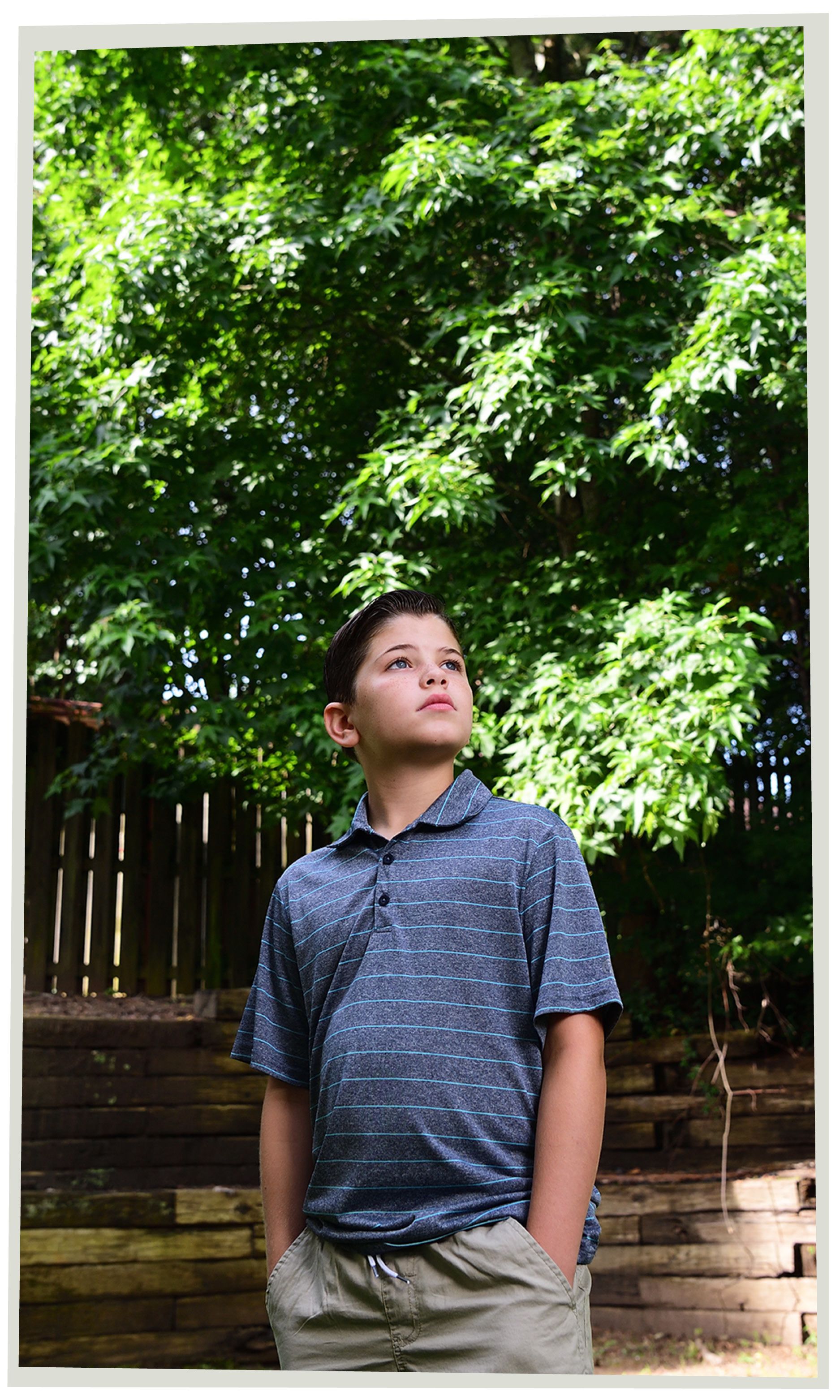 Brayden, now 10, standing in a blue shirt and tan shorts in front of the tree he fell from, a year later. 