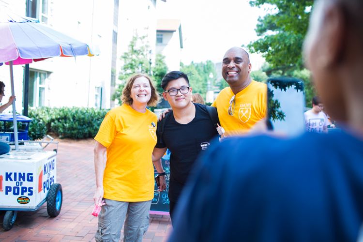 Emory President Claire E. Sterk and Provost Dwight A. McBride, wearing yellow volunteer shirts, pose with a student in a black t-shirt as someone takes their photo with a cellphone.