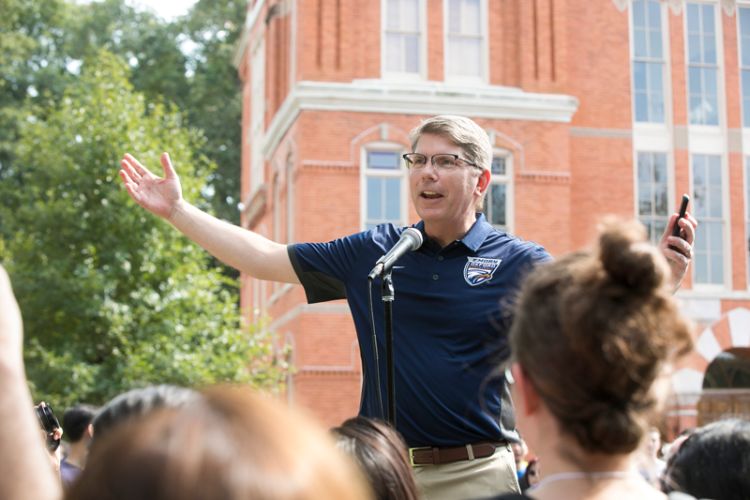 Wearing an Emory shirt, Oxford College Dean Douglas A. Hicks stands outdoors speaking to a crowd of students.