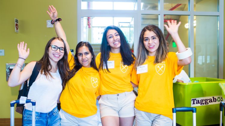 Three student volunteers in yellow shirts pose and wave with an incoming student wearing a backpack.