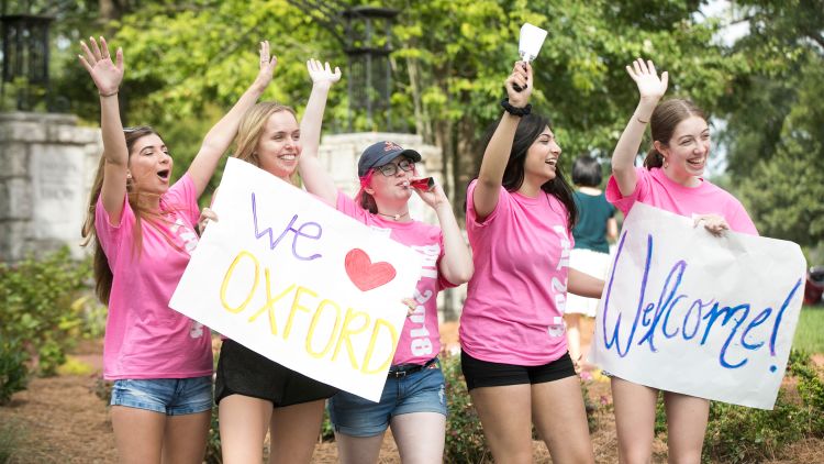 Oxford student volunteers wearing pink shirts hold signs saying "We love Oxford" and "Welcome" as they greet the Class of 2022.
