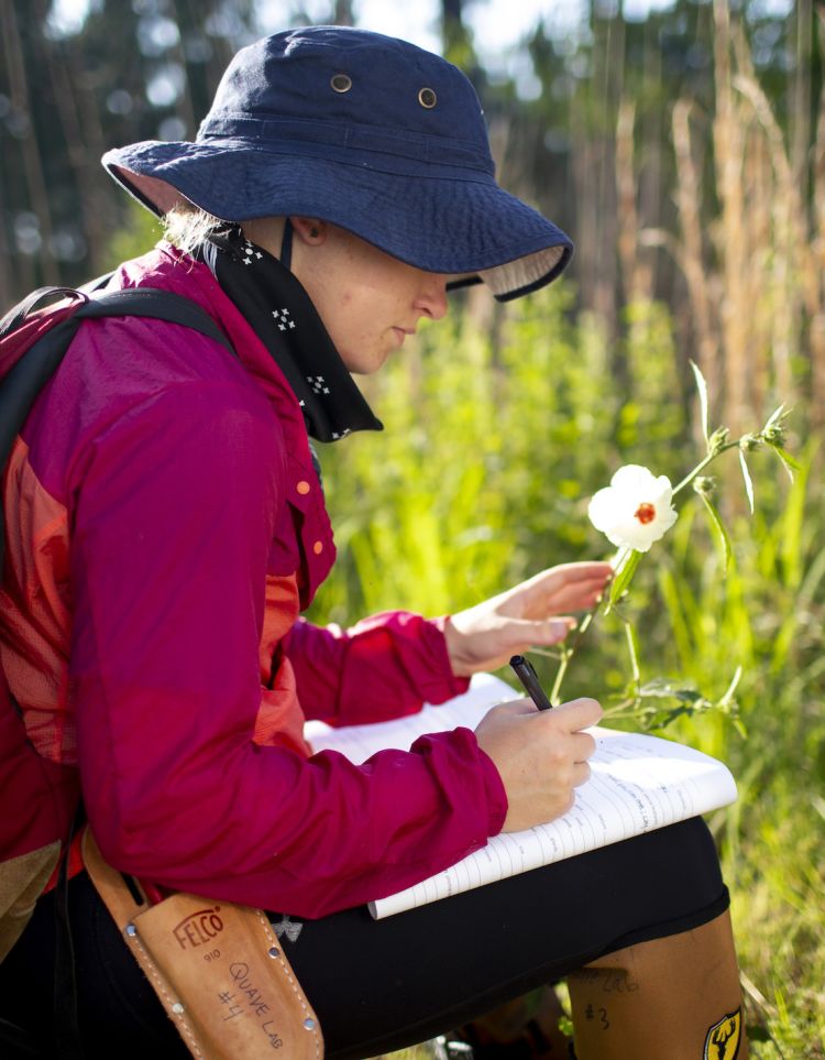 Annie Schiffer sits in the forest holding a flower and writing in a notebook