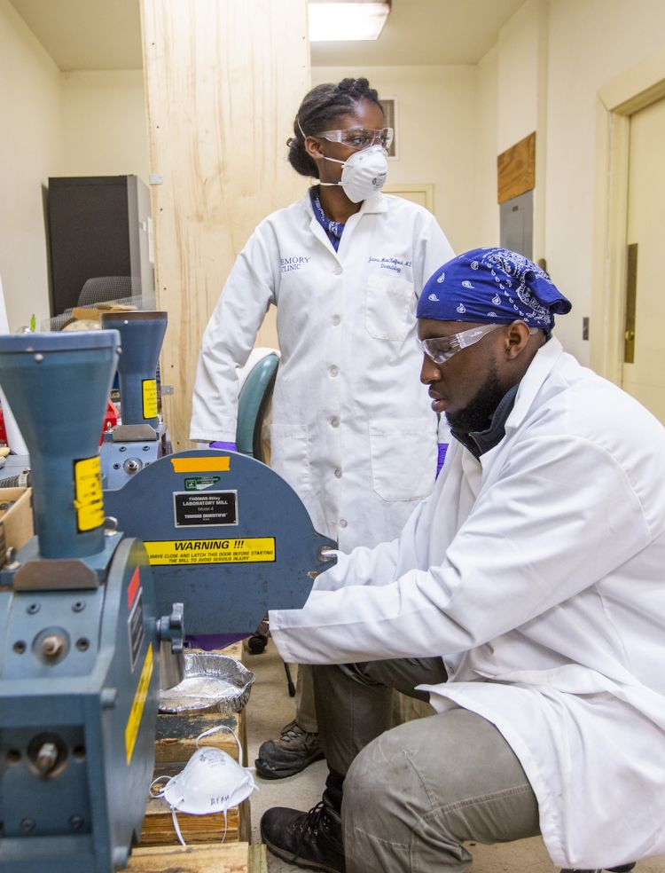 Wearing white lab coats and protective eyeware, students Kat Bagger And Afam Maduka place plants in a grinding machine