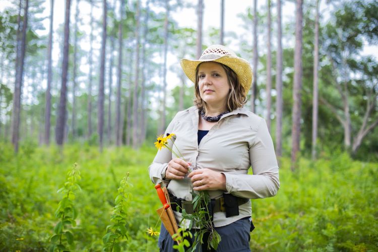 Cassandra Quave stands in the forest holding a flower and wearing a straw hat, jeans and a belt with a knife and others tools
