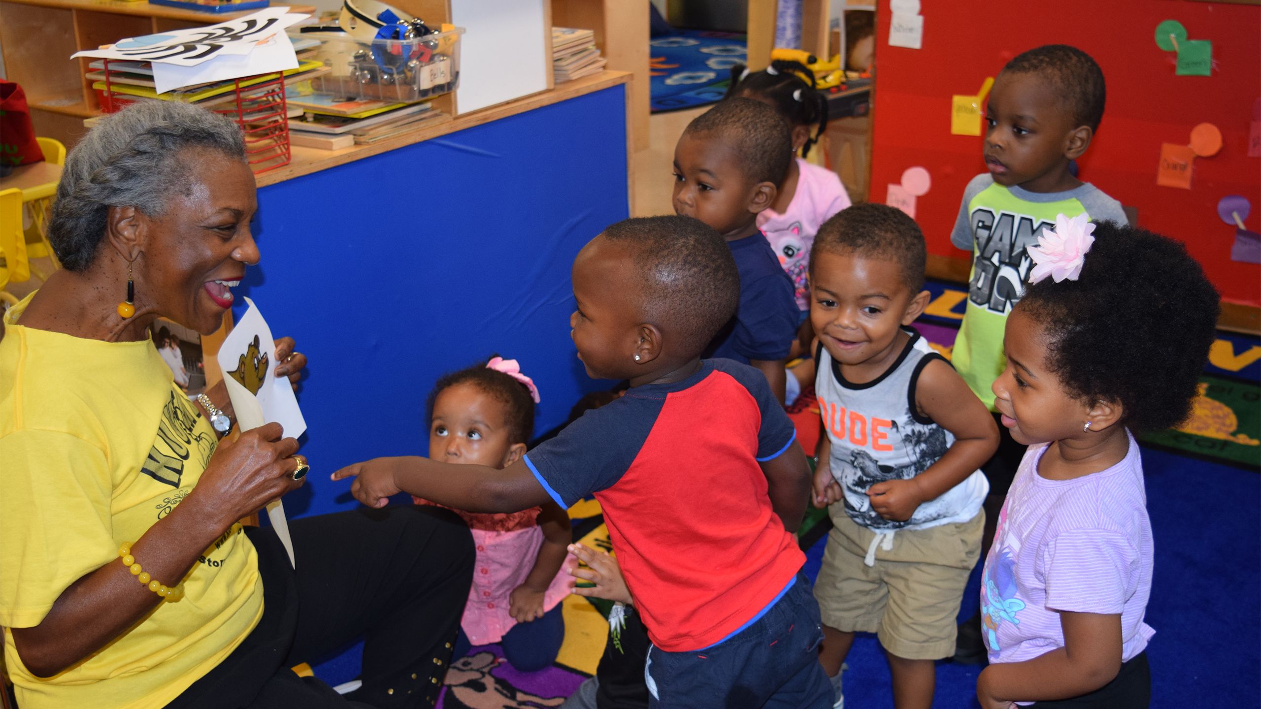 A woman holds a picture as she interacts with young children in a classroom.