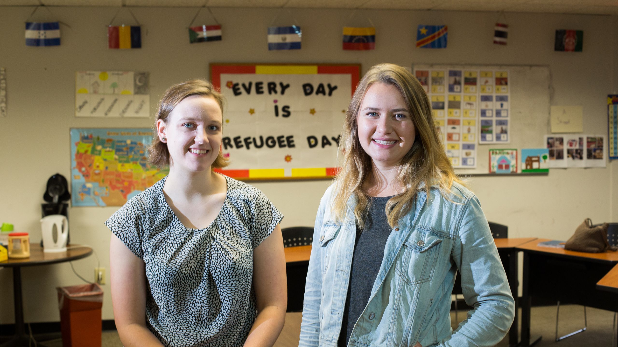 Lydia Bailey and M.K. Healy stand in a classroom with a bulletin board that reads "Every Day is Refugee Day."