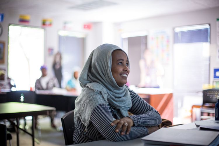 A woman with her head covered with a scarf smiles as she sits at a table in a classroom at the International Rescue Committee.