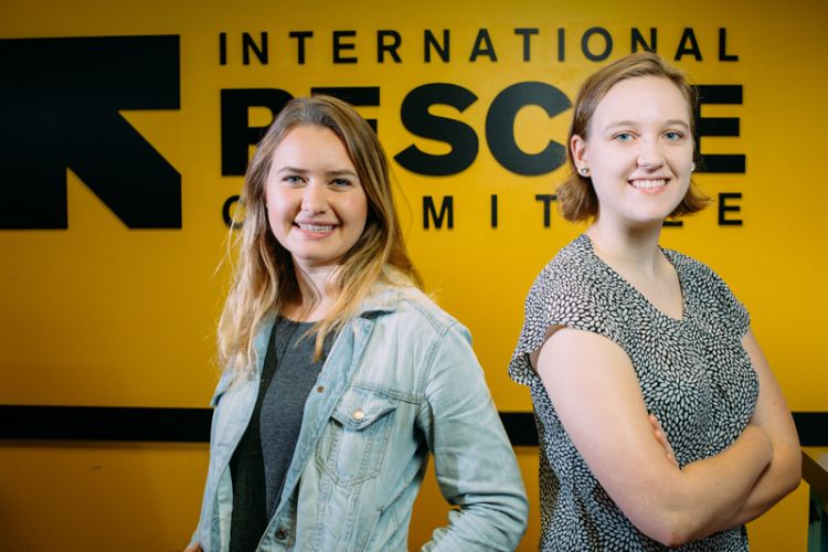 M.K. Healy and Lydia Bailey stand in front of a yellow sign that shows the logo and name of the International Rescue Committee.