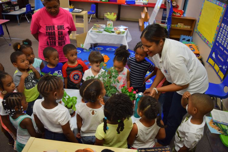 Young children surround a table and listen as a smiling woman shows them plants.