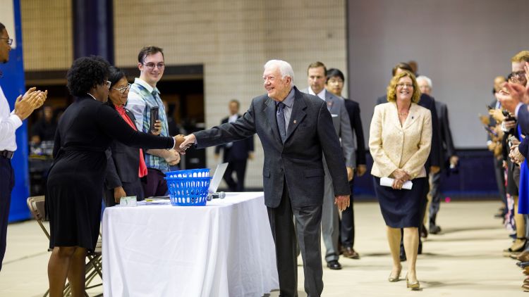 President Jimmy Carter shakes hands with a woman as he enters Emory's Woodruff PE Center for the 37th annual Carter Town Hall. Emory President Claire E. Sterk and others walk behind him as the audience claps.
