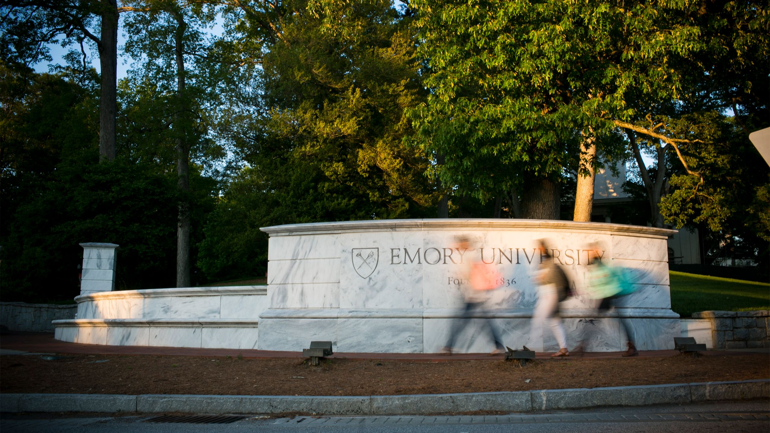 Students walk in front of the white wall that says "Emory University" at the main gate to campus