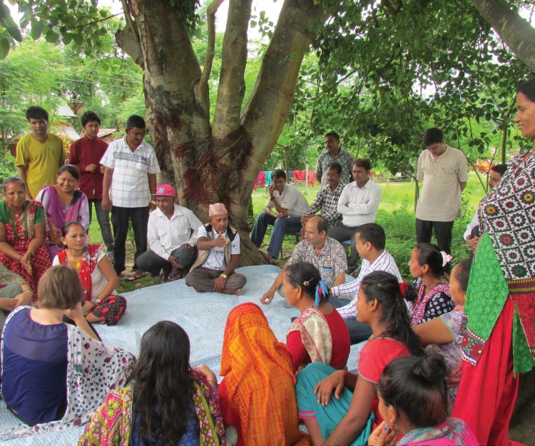 A group of men and women in traditional Nepali garb sit in a large circle under a tree.