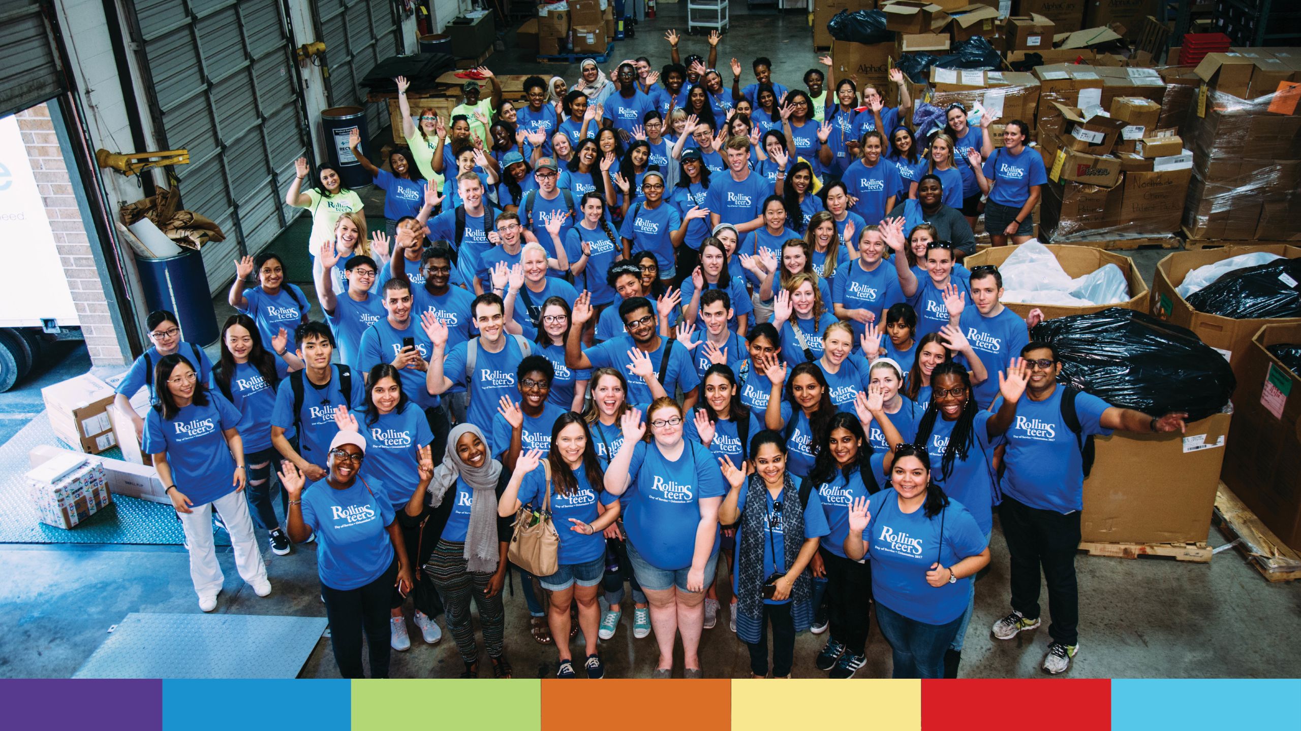 A large group of students in blue Rollins-teer T-shirts wave at the camer