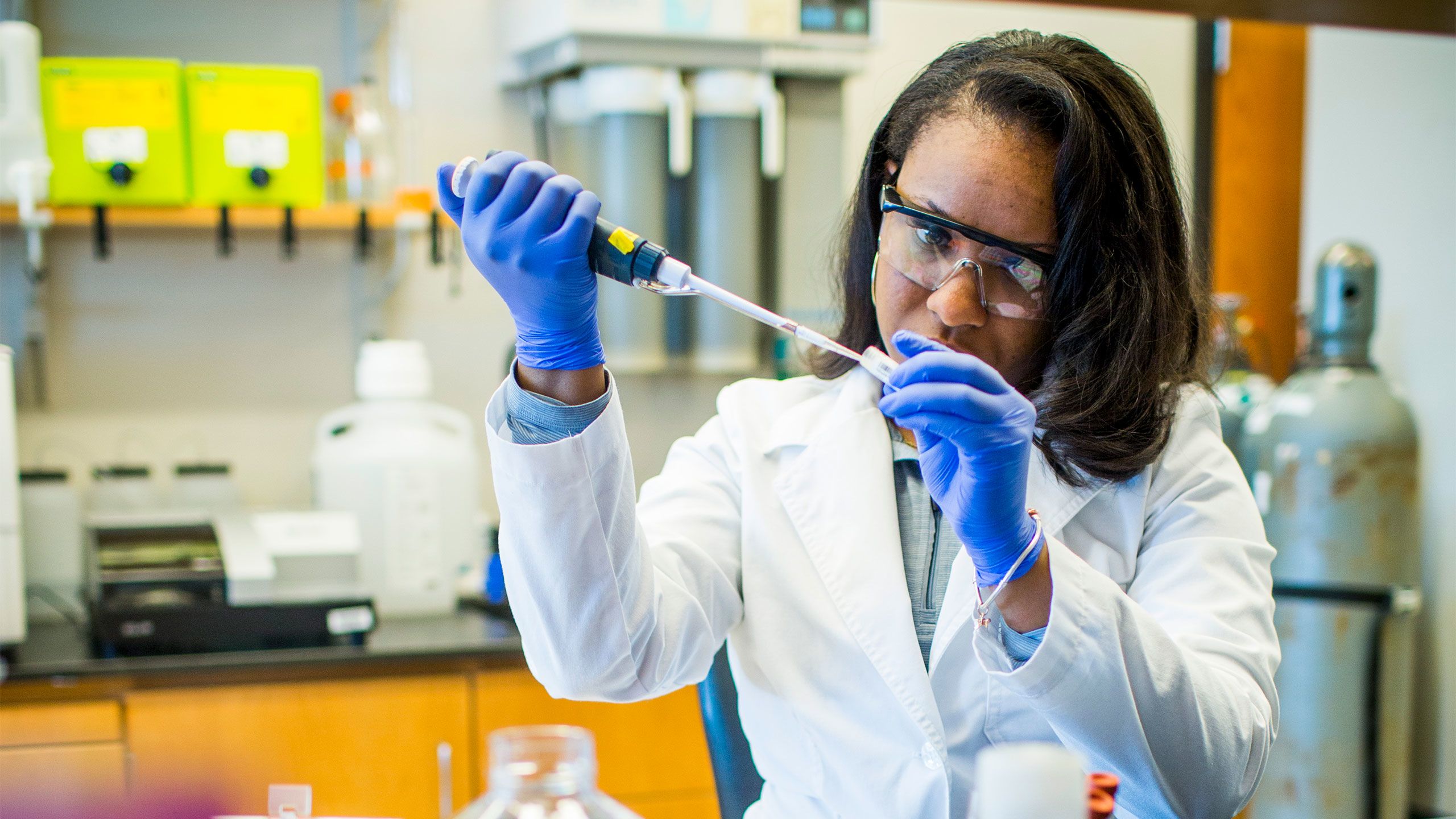 A woman wearing a white coat, gloves and protective eye wear works in a lab.