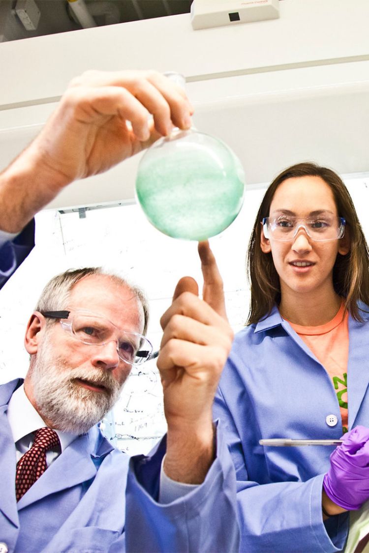 A professor holds up a beaker as a student looks on. Both are wearing blue labcoats and protective eyewear. 