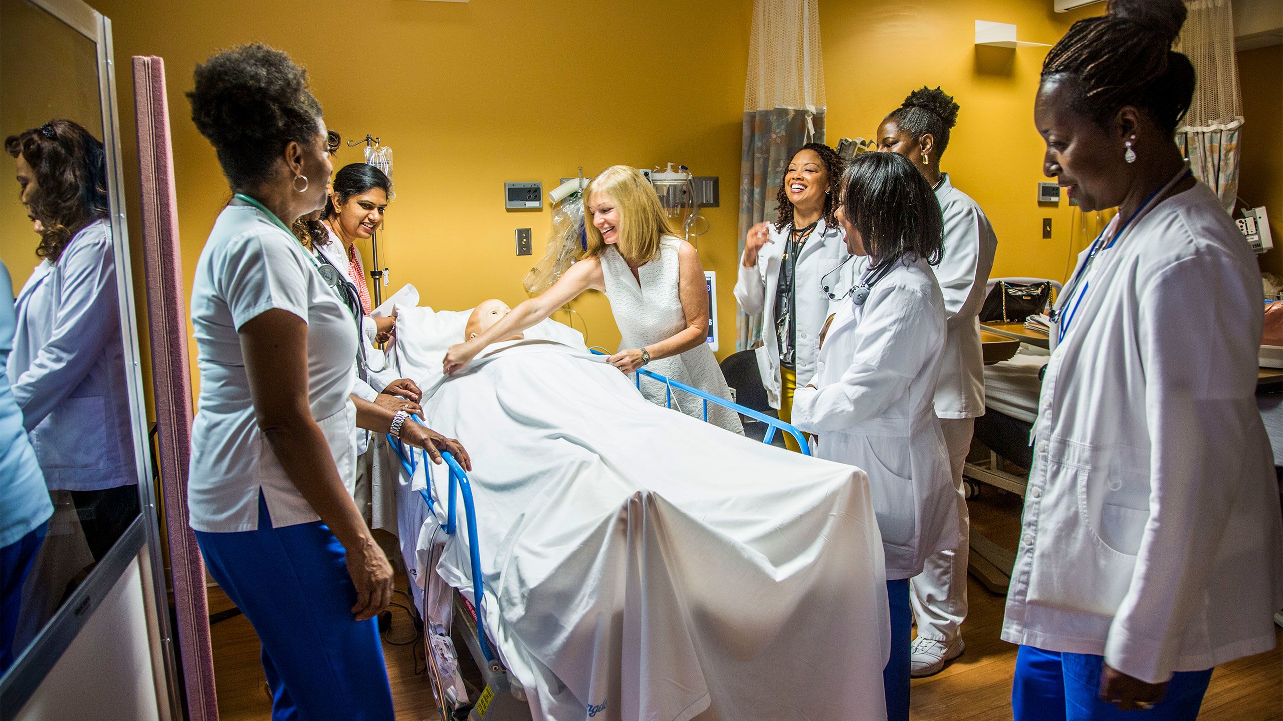 Nurses wearing white coats practice with a plastic simulated patient lying on a hosital bed.
