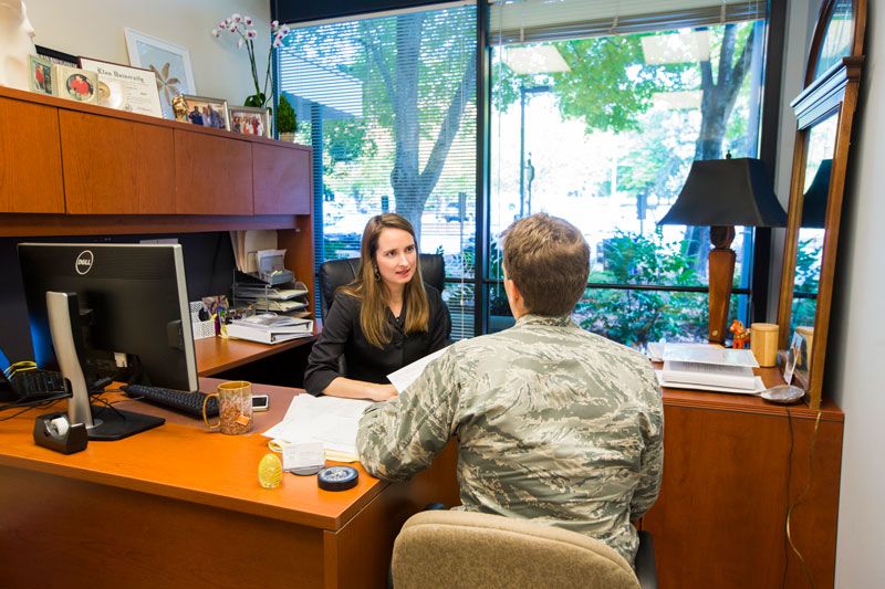 Keely Youngblood sits at a desk facing the camera; a man in a military uniform is talking with her with his back to the camera.