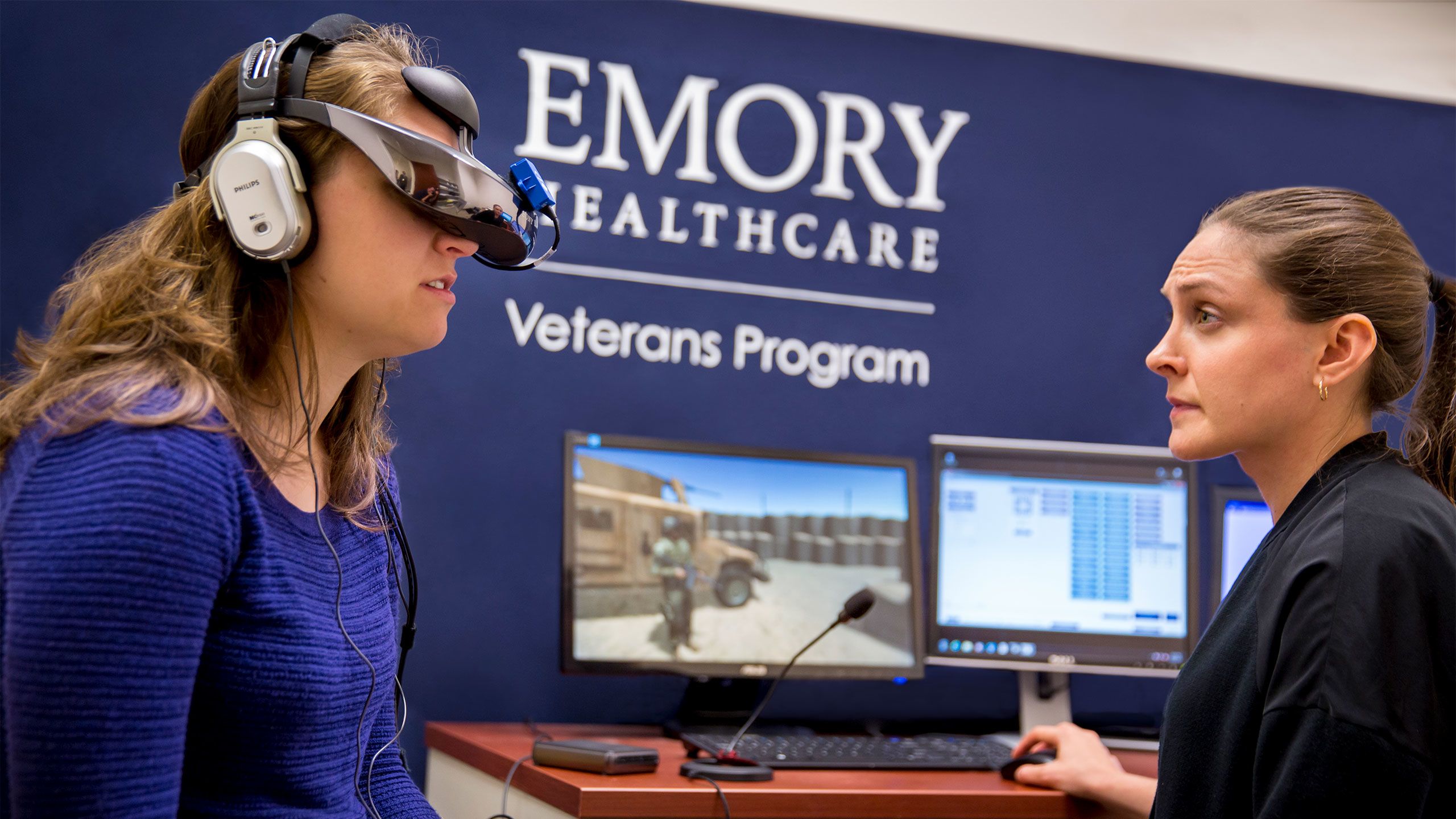 A woman wears a virtual reality headset. Computer screens show what she sees: a military truck. 