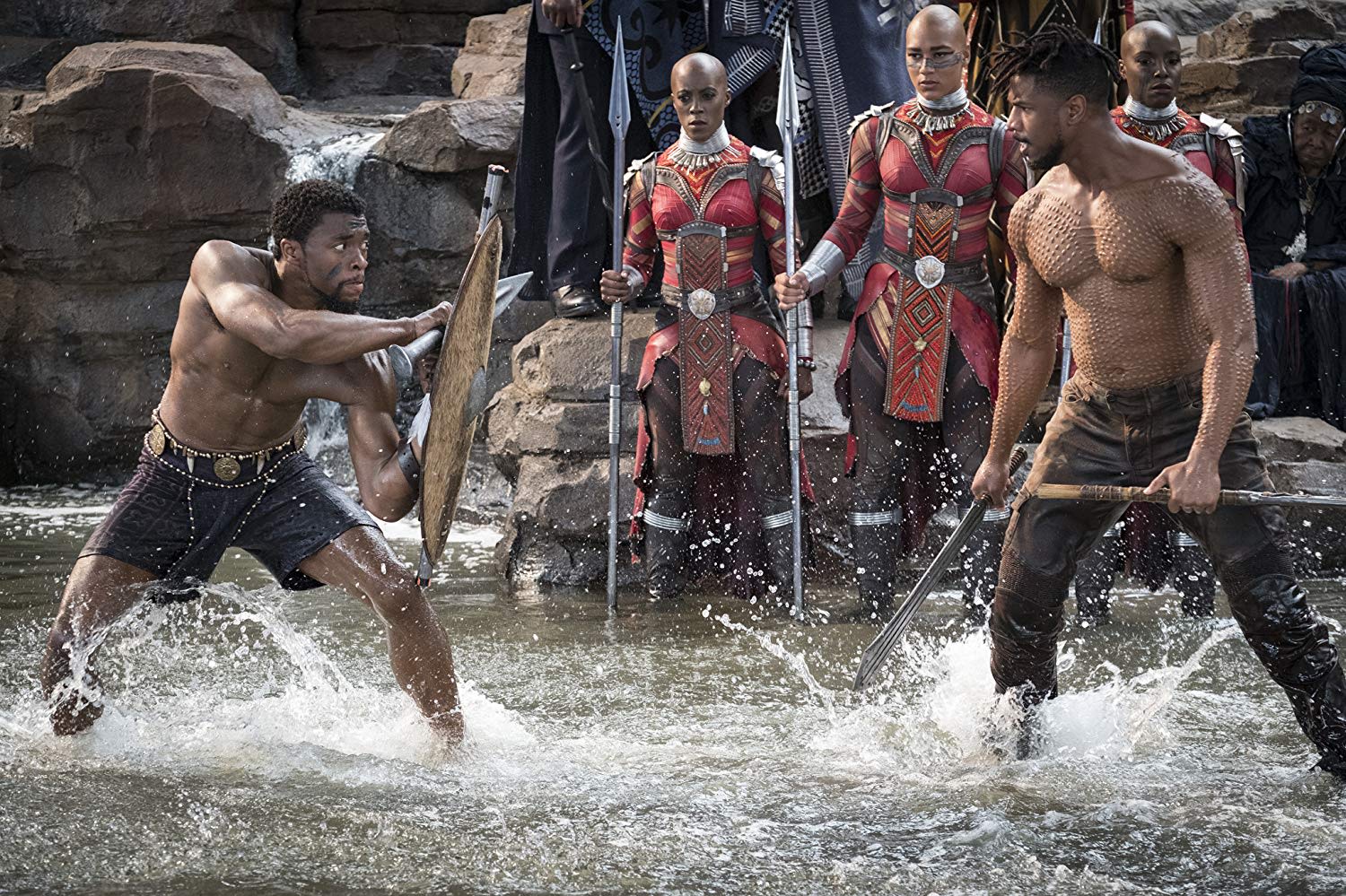 Actors Chadwick Boseman and Michael B. Jordan from the film "Black Panther" stand in shallow water, holding weapons, and prepare to fight. Three women look on.