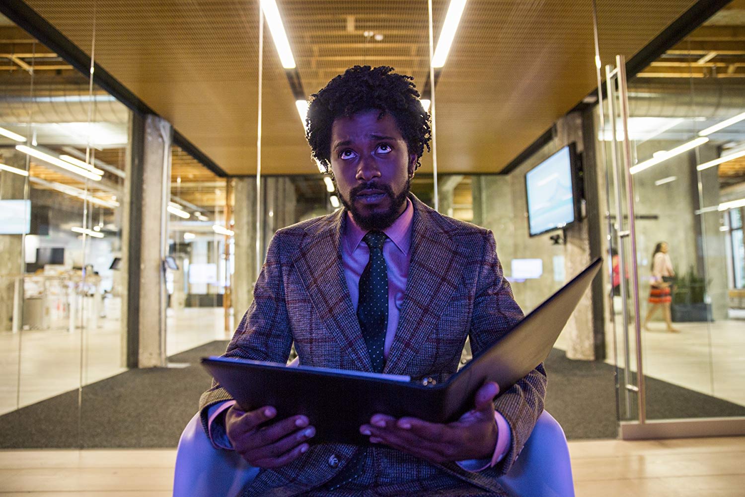 Actor Lakeith Stanfeld wears a plaid suit jacket and polka dot tie while holding open a binder and sitting outside of an office building in the film "Sorry to Bother You."