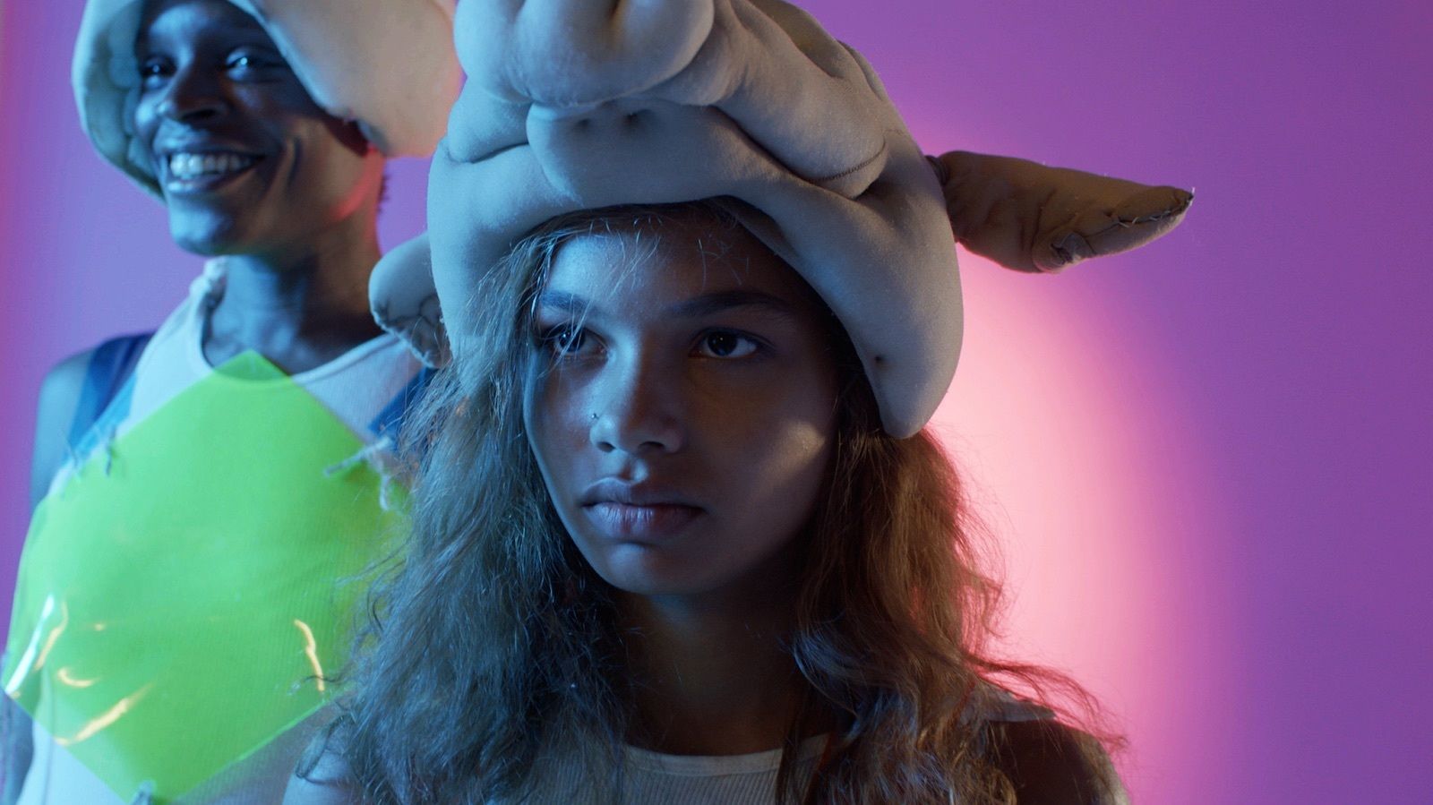 Actress Helena Howard wears an animal head in the film "Madeline's Madeline." A young man stands behind her in another animal head. Bright purple background. 