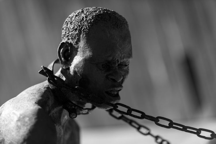 An up-close photo shows a statue of an enslaved person with a chain around his neck.