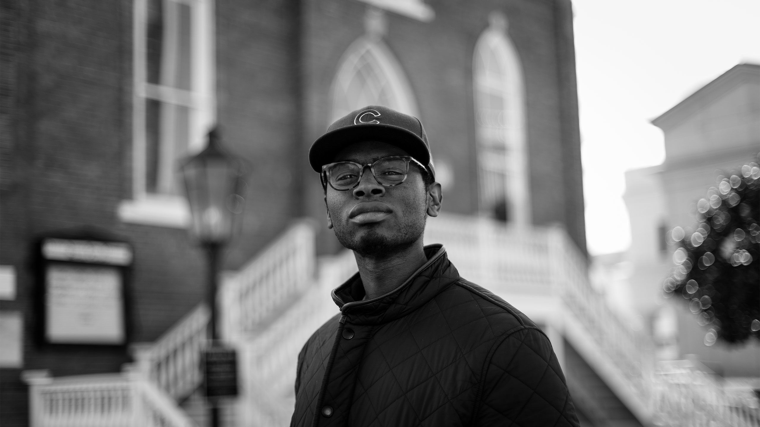 A portrait shows Julian Reid wearing glasses and a baseball cap standing outside of a church.