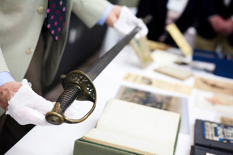 Burkett wearing white gloves to hold a sword out over a table upon which other archival materials are on display