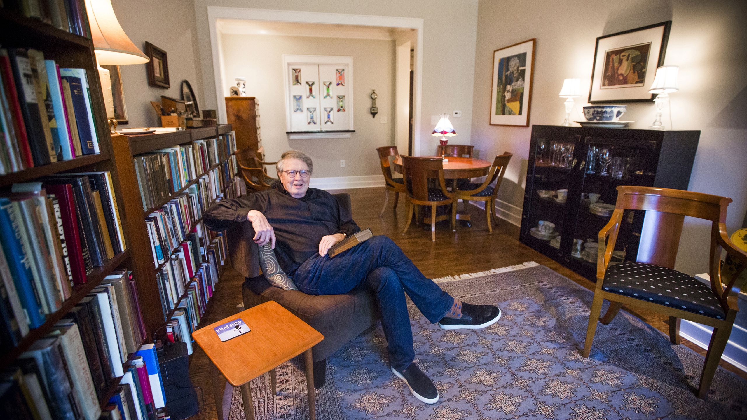 Burkett smiling and leaning back in a chair in his home library with his legs crossed and a book resting in his lap
