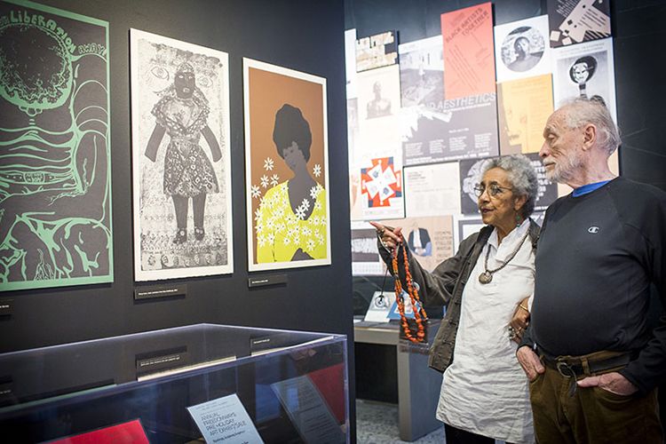 Camille Billops and James Hatch standing close to one another, arms hooked, looking at materials on display in the gallery. Camille is pointing with a hand holding a beaded necklace.